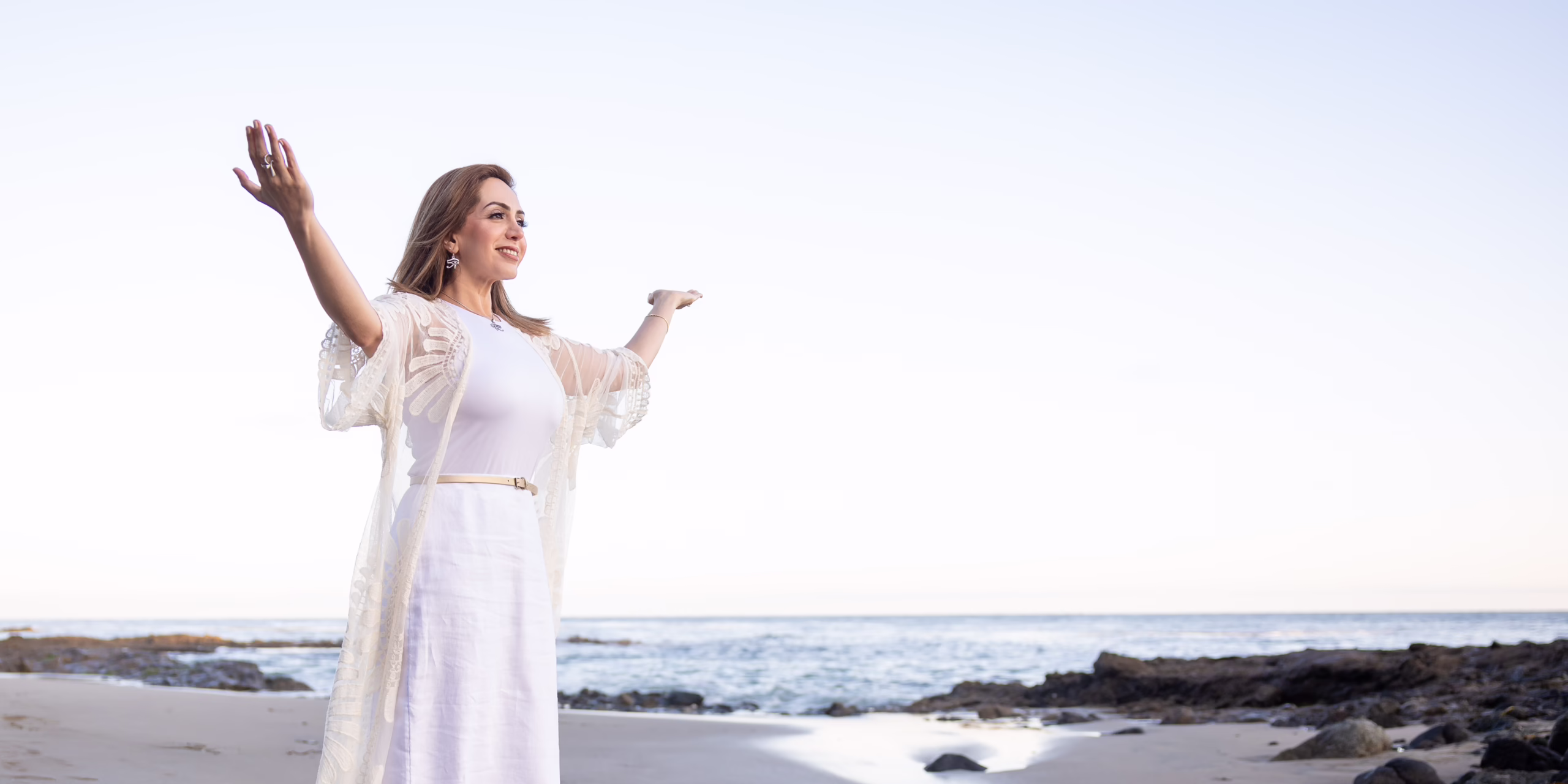 A woman in white with arms raised at the ocean's edge